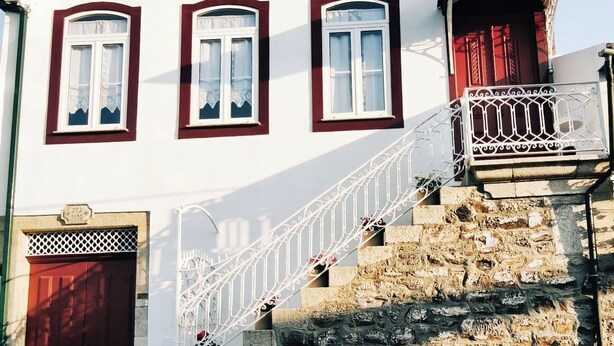 A white building with three arched windows, a red door, and a stone staircase leading to a balcony with a white railing.