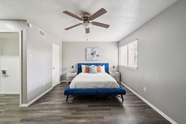 A bedroom with a bed, a ceiling fan, a nightstand, a window with blinds, and a door.
