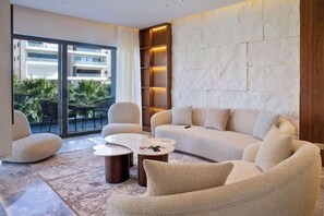 Un salon moderne avec un canapé incurvé, deux fauteuils et une table basse. Une grande fenêtre offre une vue sur des palmiers et un immeuble à l’extérieur.