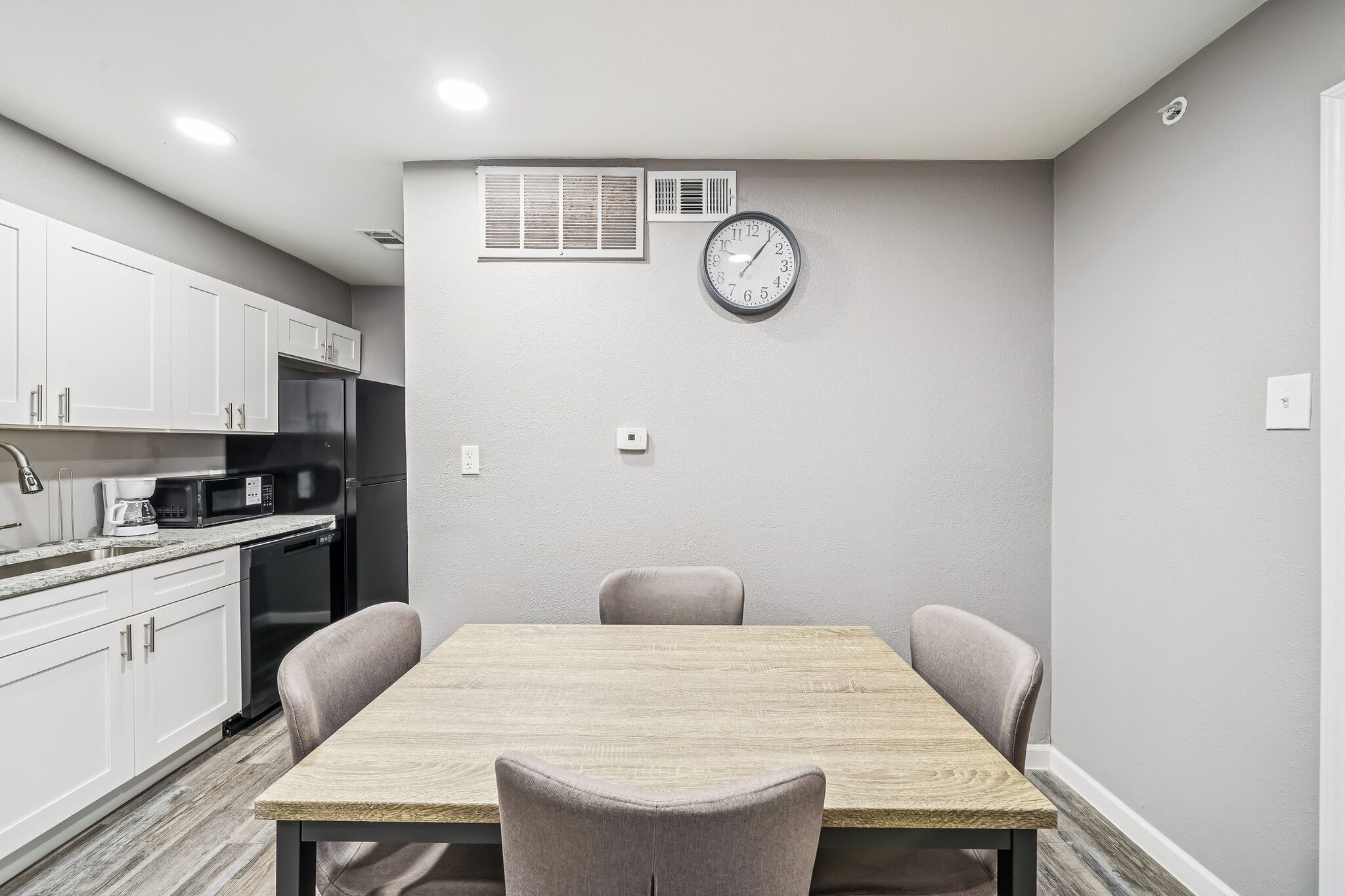 A modern kitchen with white cabinets, a microwave, and a refrigerator. There is a dining table with four chairs and a clock on the wall.