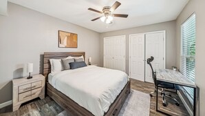 Ein Schlafzimmer mit Bett, Nachttisch, Schreibtisch, Stuhl, Deckenventilator und zwei Türen.