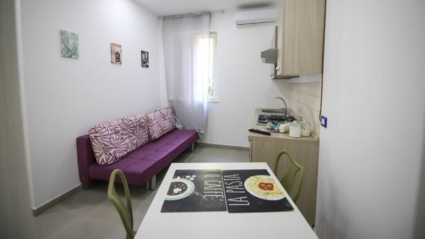 A small, well-lit room with a purple sofa, a dining table with placemats, a kitchenette with a sink and cabinets, and a window with a curtain. - Residenza Cicerone (Agrigento)