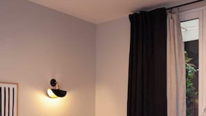 A hotel room with a bed, a wall-mounted light fixture, a window with curtains, and a view of greenery outside. - Hotel Tinah: Elegance and Comfort Near the Champs-elysees (Paris)