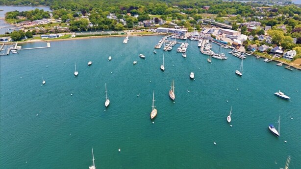 An aerial view of a marina with numerous boats docked, surrounded by a residential area and a forested landscape.
