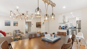 A modern living room with a large wooden dining table, a kitchen in the background, and a chandelier with multiple light bulbs.
