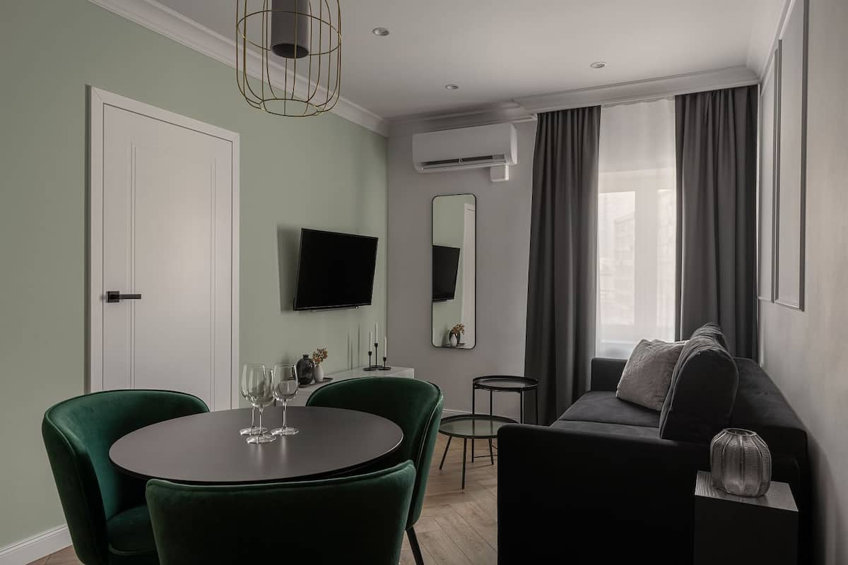 A modern living room with a round table, two green chairs, a grey sofa, a flat-screen TV, a mirror, and a window with curtains.