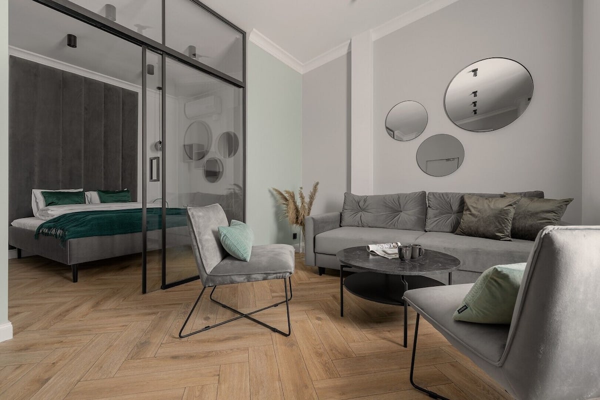 A modern living room with a sofa, armchair, and coffee table. A bed with a green blanket is visible through a glass door.