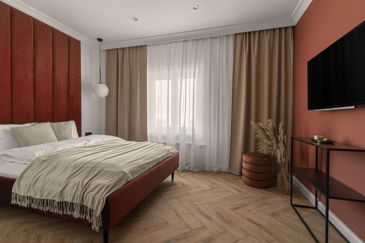 A modern hotel room with a bed, a TV, a wooden floor, and a large window with curtains.