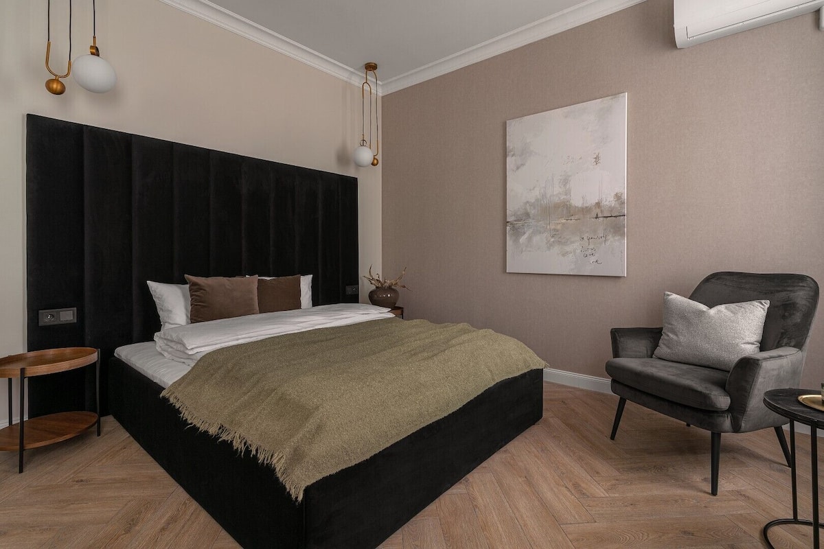 A modern bedroom with a large bed, a dark headboard, a grey armchair, and a minimalist decor style.