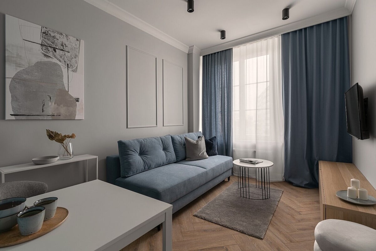 A modern living room with a grey sofa, a coffee table, a TV mounted on the wall, and a large window with blue curtains.