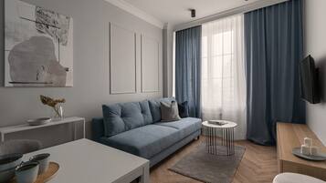 A modern living room with a grey sofa, a coffee table, a TV mounted on the wall, and a large window with blue curtains.
