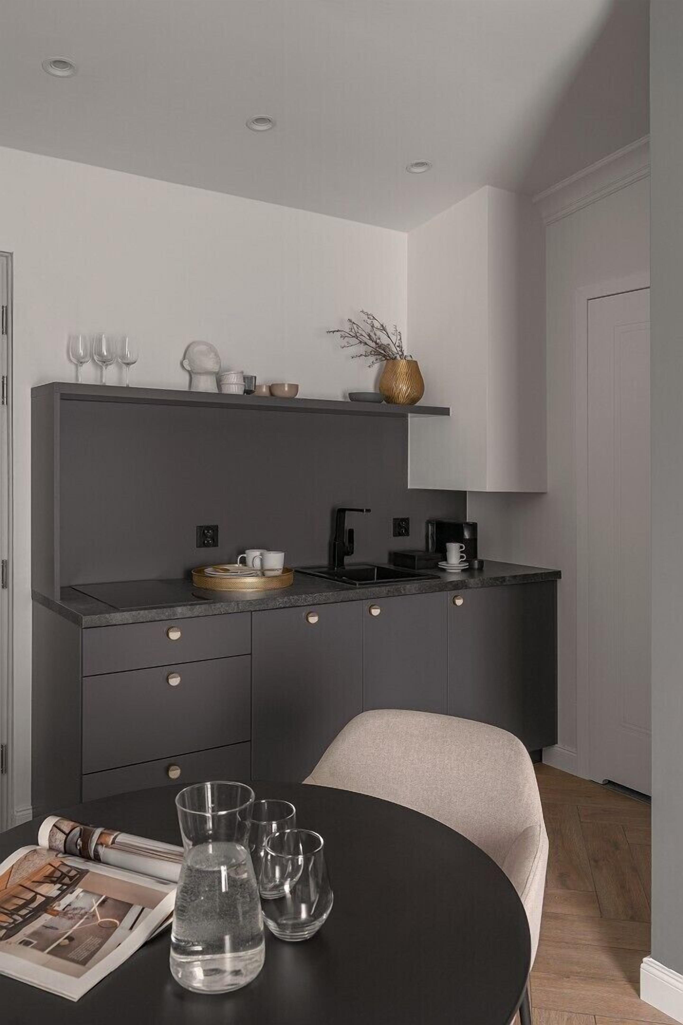 Una cocina moderna con encimera oscura, un lavamanos y una cafetera. Hay una mesa de comedor con una revista y vasos.