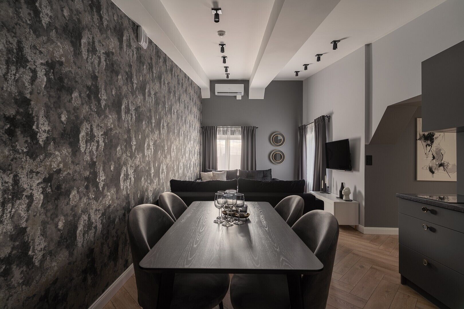 Un comedor moderno con una mesa y sillas de madera, un sofá oscuro y un televisor de pantalla plana montado en la pared.