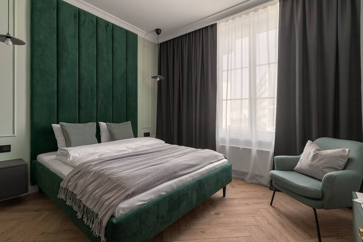 A bedroom with a large bed, a green armchair, and a window with sheer curtains.