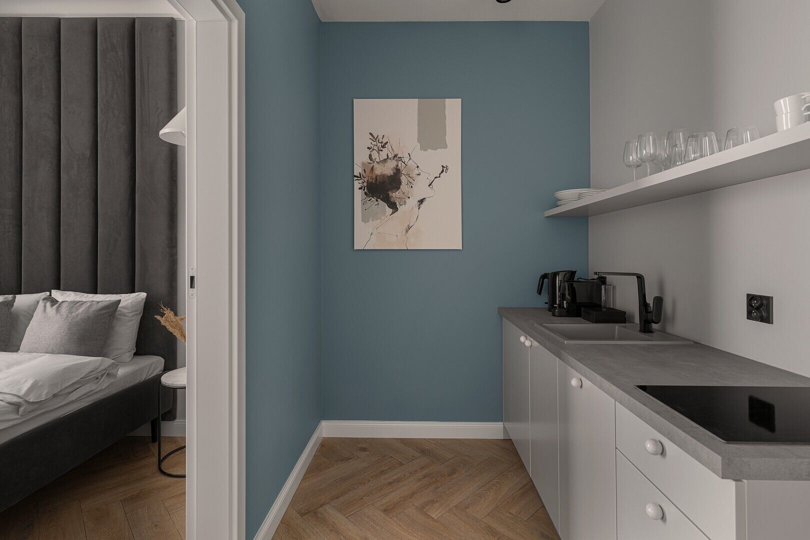 A modern bedroom with a bed, bedside table, and a wall-mounted lamp. Adjacent to the bedroom is a kitchen with a countertop, sink, and a coffee machine.