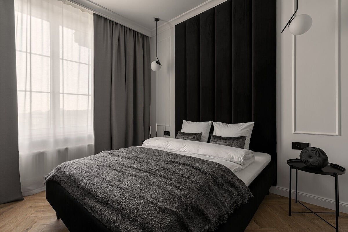 A modern bedroom with a large bed, a bedside table, a lamp, and a view of the outside through a window with curtains.