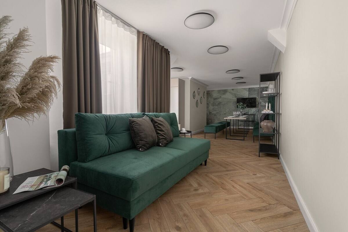 A modern living room with a green sofa, a small table, and a dining area in the background.