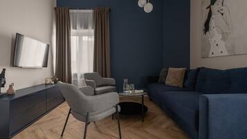 Una sala moderna con un sofá azul oscuro, un sillón gris, una mesita de centro redonda y un televisor de pantalla plana montado en la pared.