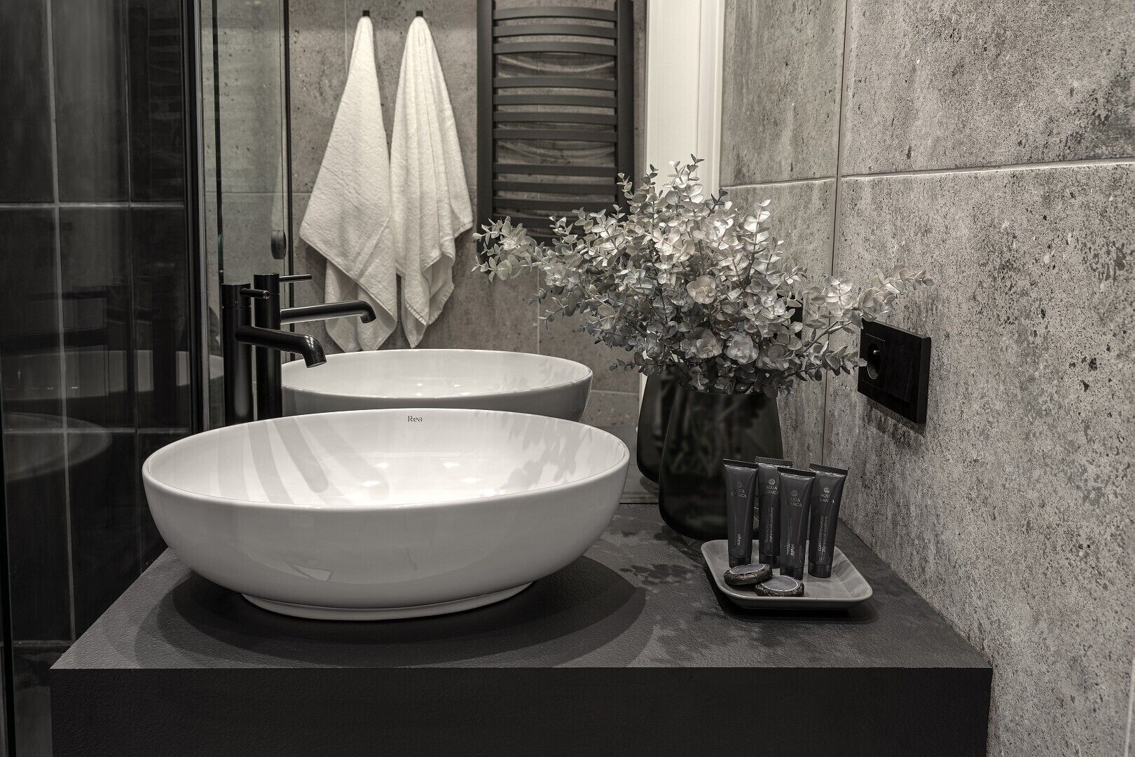 Un baño moderno con dos lavamanos grandes blancos, cabina de ducha de vidrio y un florero con flores en la encimera.