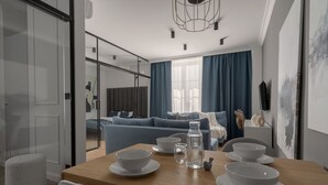 A modern living room with a sofa, a dining table with bowls and glasses, and a large window with curtains. - Apartio Rooms Katowice (Katowice)