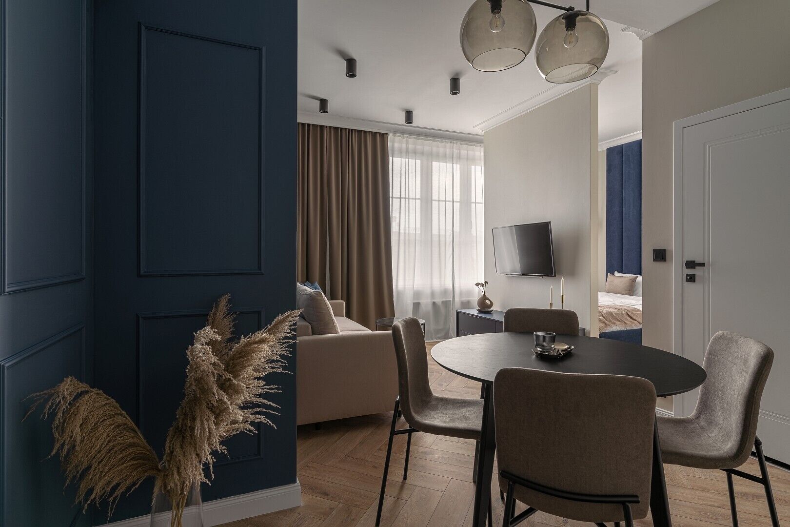 A modern hotel room with a dark blue accent wall, a round dining table with chairs, a sofa, and a TV mounted on the wall.