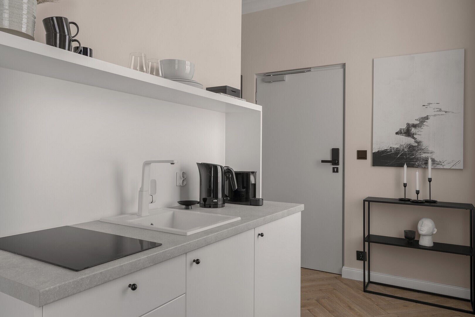 Una cocina moderna con un lavabo, hervidor y cafetera, un armario blanco y una puerta al fondo.