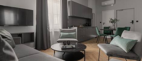 A modern living room with a sofa, armchair, and coffee table. There is a flat-screen TV mounted on the wall, a kitchen area with cabinets, and a dining table with chairs.