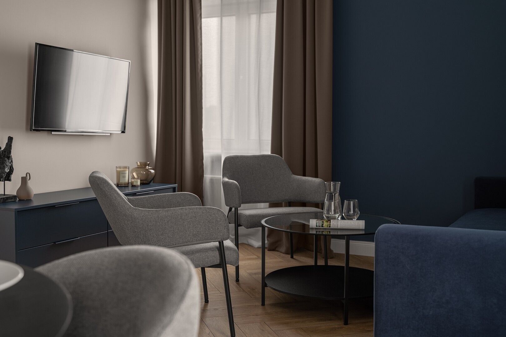 Een moderne hotelkamer met een flatscreen-tv, twee grijze fauteuils, een ronde salontafel en een blauwe bank.