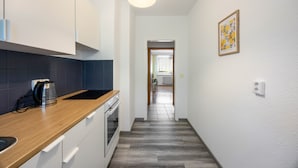 Un couloir étroit, avec un coin cuisine sur la gauche, un plancher en bois et une porte donnant sur une autre pièce.