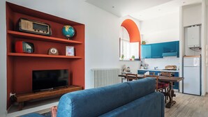 Un salon moderne avec un meuble de rangement rouge pour l’équipement audiovisuel, un canapé bleu et une cuisine dotée de meubles bleus et d’un réfrigérateur blanc.