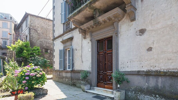 Un bâtiment historique doté d’une grande porte en bois, d’un balcon orné de fleurs et d’un petit jardin où poussent des fleurs roses.