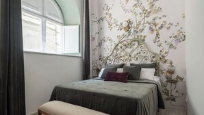Une chambre à coucher avec un lit, un banc, une fenêtre avec des rideaux et un mur orné d’un motif floral.