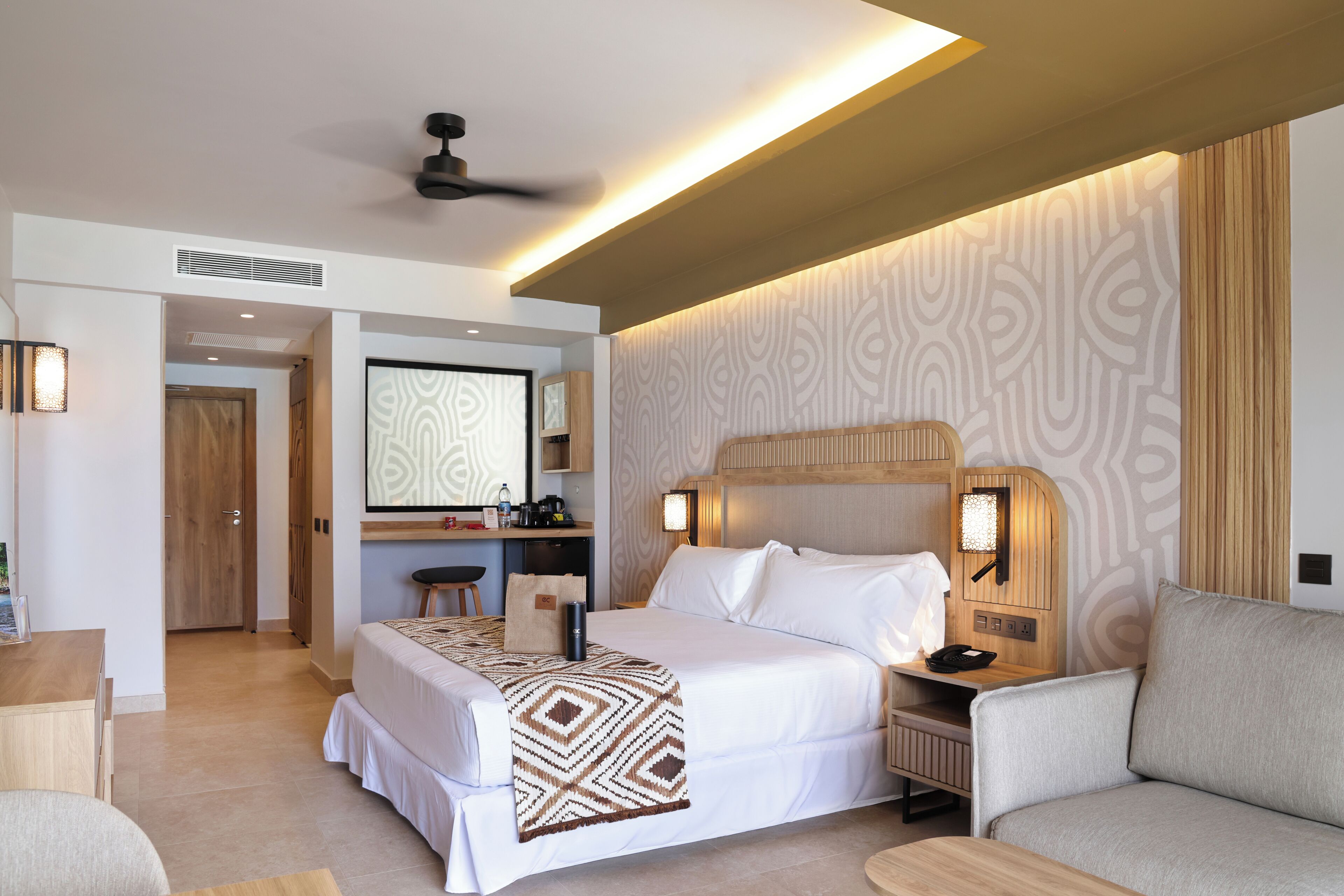 Une chambre d’hôtel équipée d’un grand lit, d’un bureau, d’une chaise, d’un canapé, d’un ventilateur de plafond et d’un téléviseur fixé au mur.