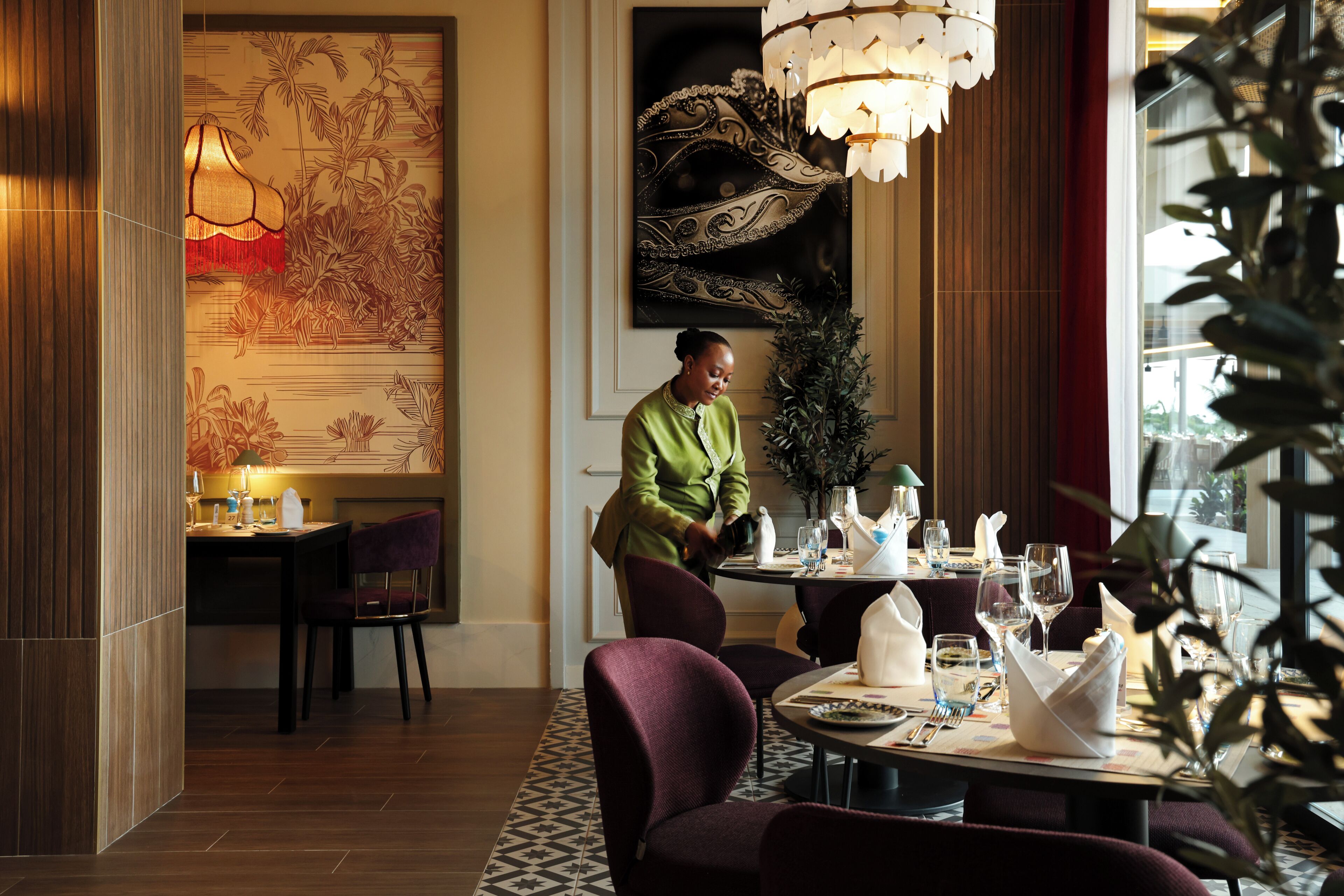 A dining area with a table set for two, a chandelier, and a large decorative wall piece.