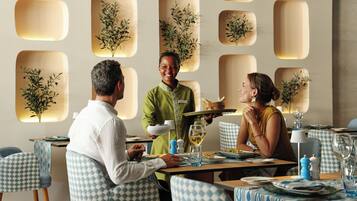 Un restaurante con sillas a cuadros, un camarero atendiendo y una mesa con copas de vino.