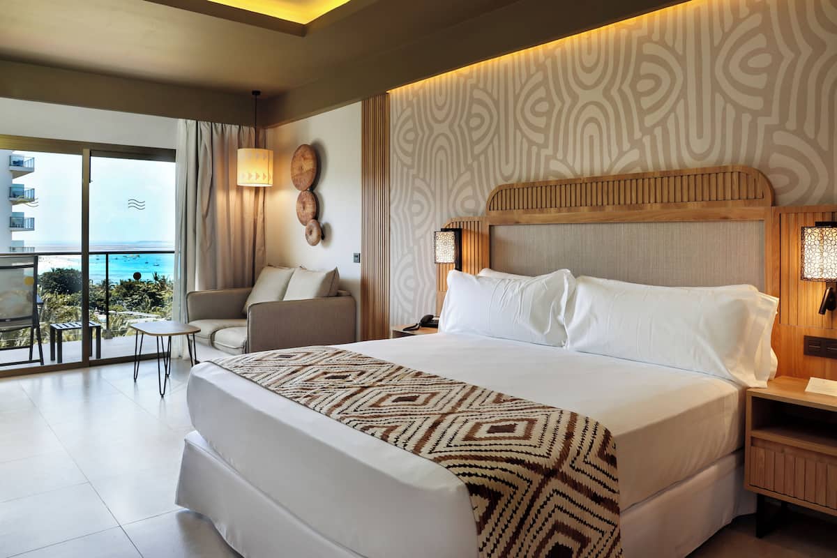 A hotel room with a large bed, a chair, a small table, and a view of the ocean.