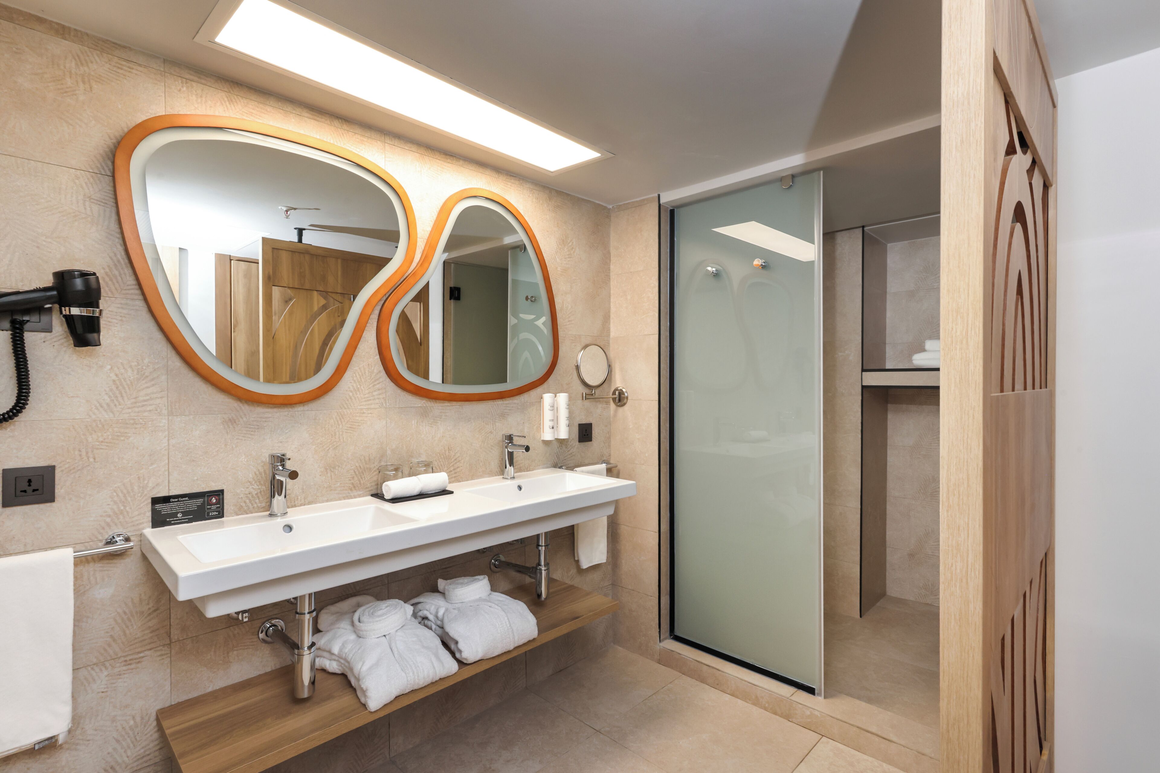 A modern bathroom with a double sink, a mirror with an orange frame, a shower area, and a wooden vanity with towels.