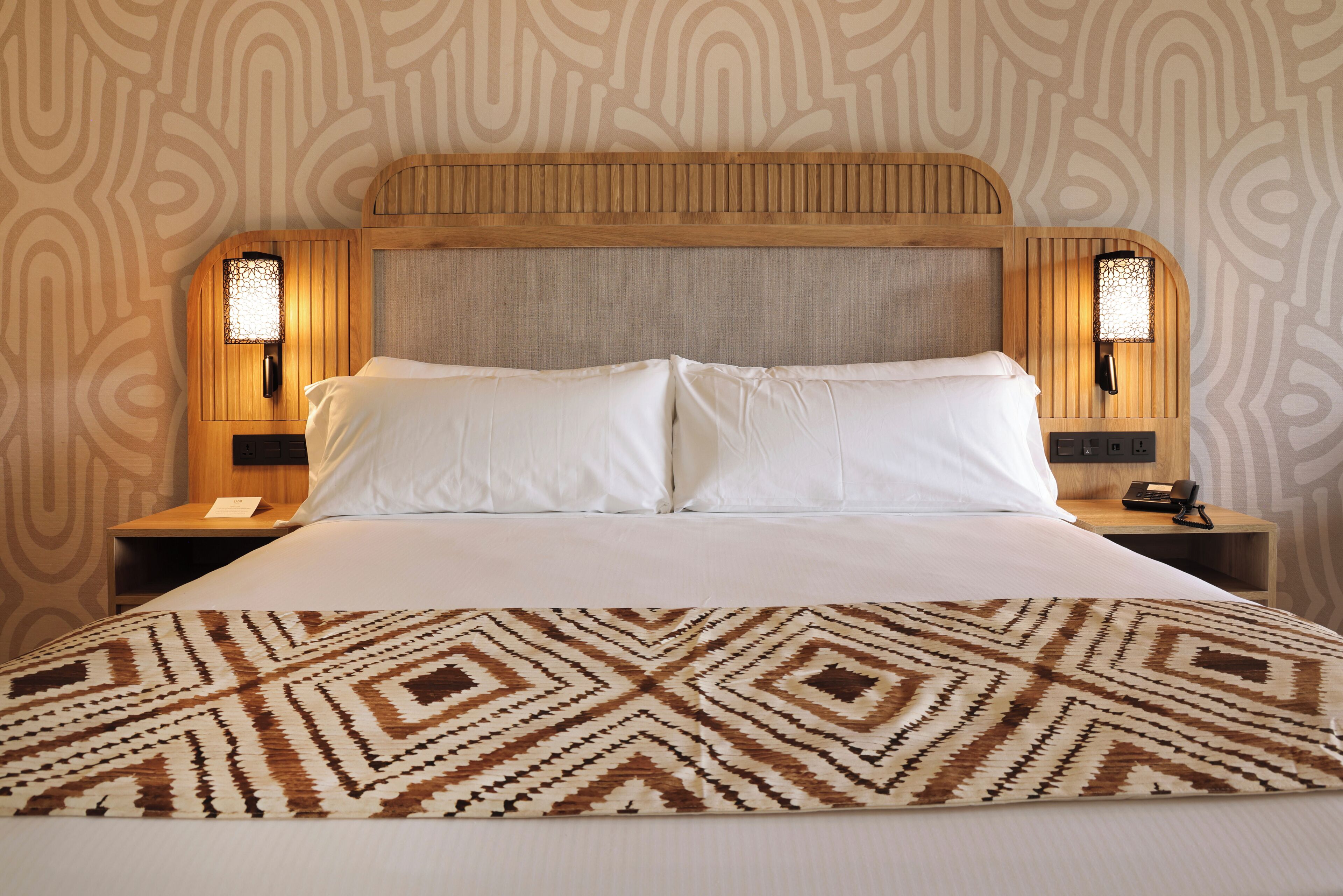 Una habitación de hotel con una cama grande, dos mesitas de noche con lámparas y un papel tapiz estampado.