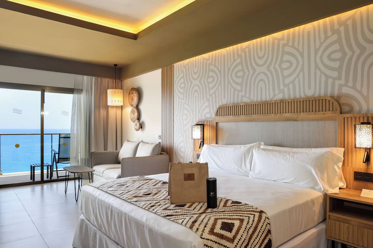 A hotel room with a bed, a bedside table, a chair, a small table, and a view of the ocean.