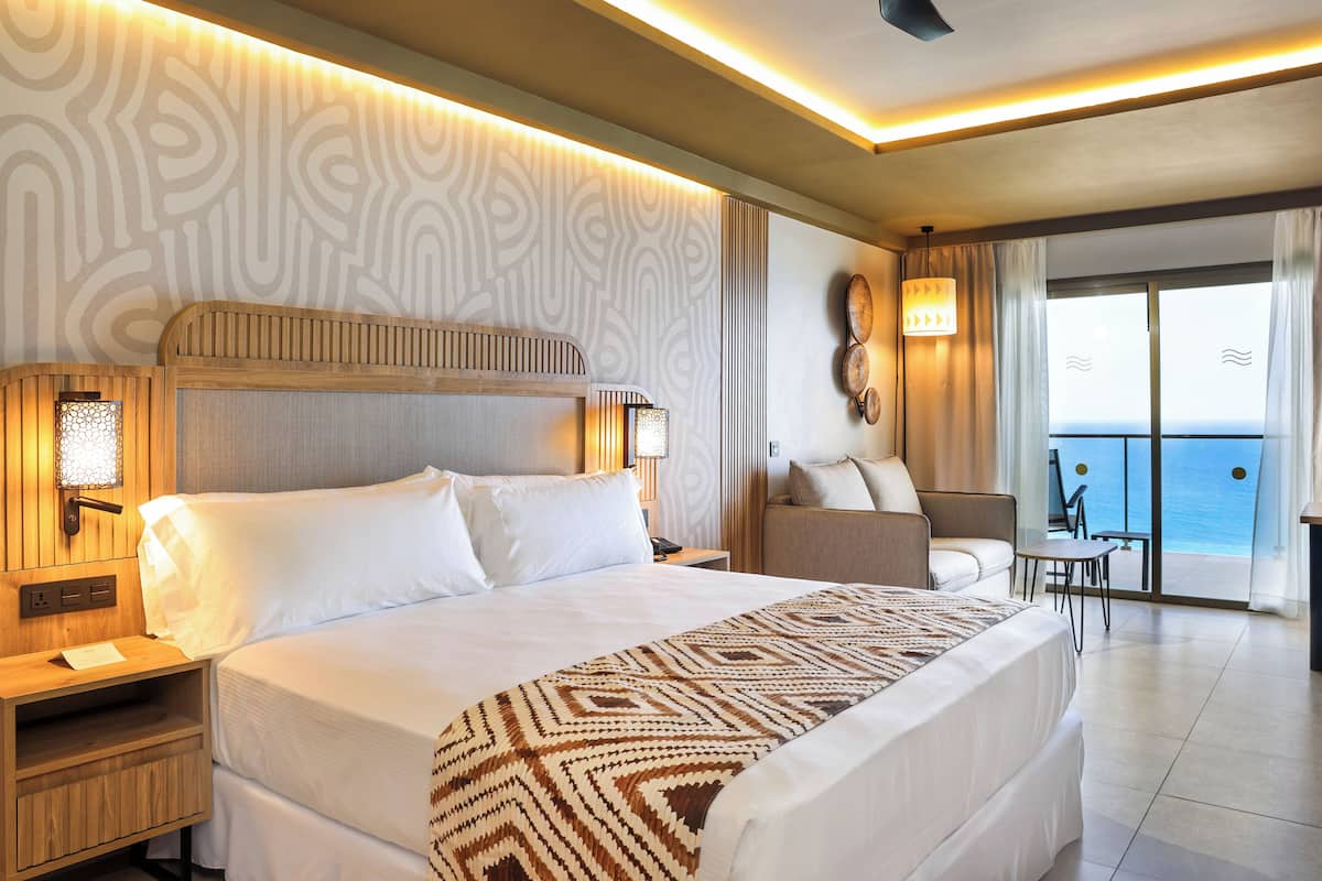 A hotel room with a large bed, a nightstand, a desk, a chair, and a view of the sea through a sliding door.