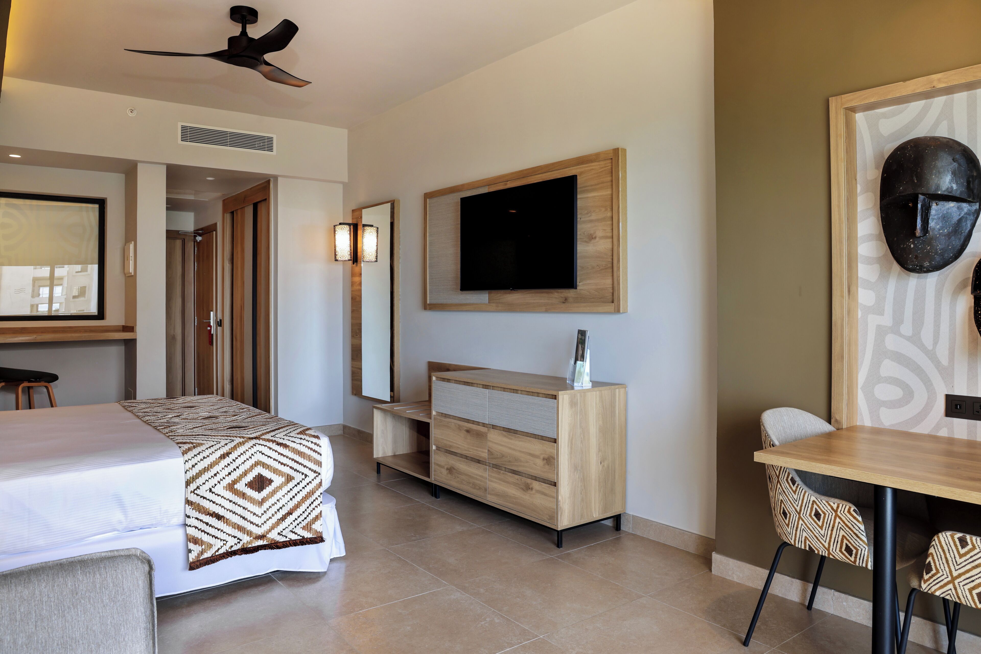 Ein Hotelzimmer mit Bett, Schreibtisch, Stuhl, Wandfernseher und Deckenventilator.