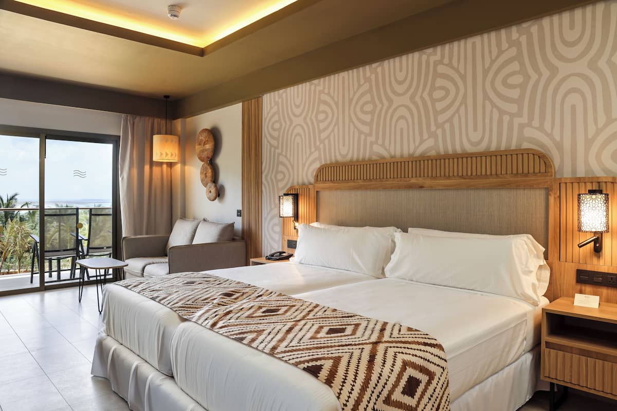 A hotel room with a large bed, a sofa, a small table, and a balcony with a view.