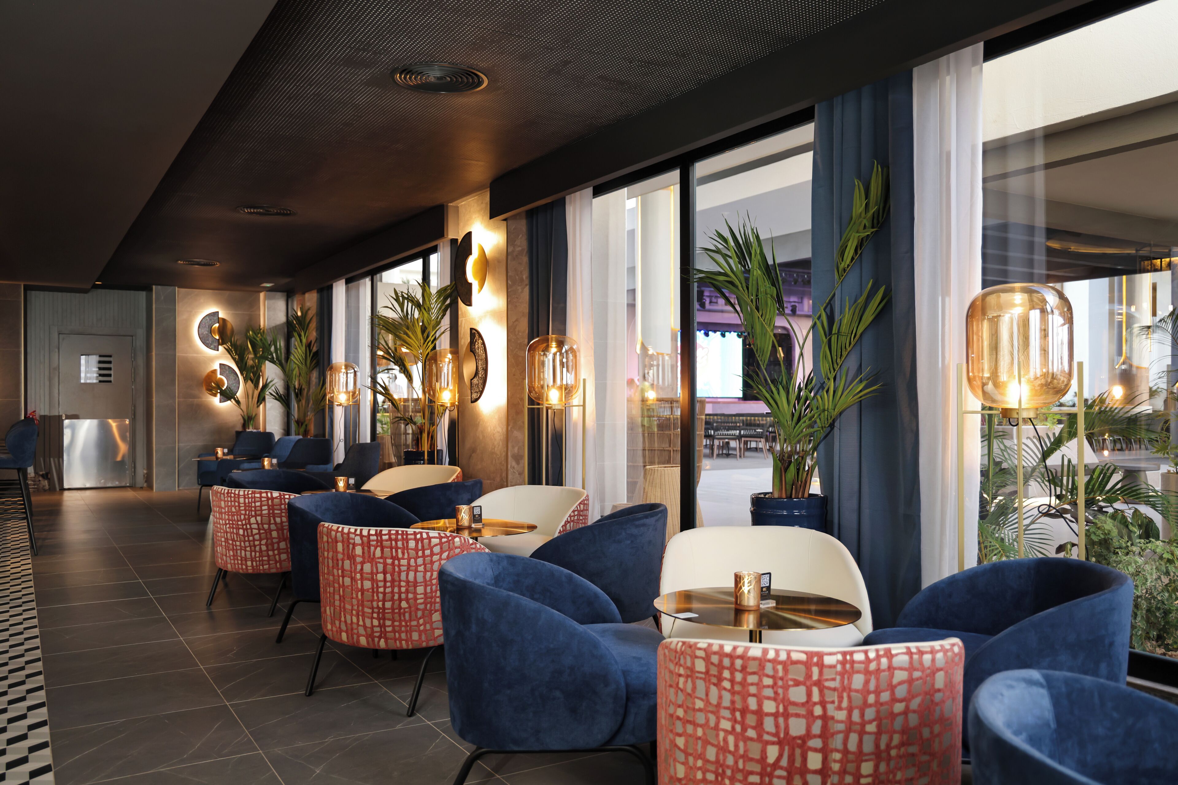 Un hall d’hôtel moderne, doté d’espaces aménagés avec des sièges, de grandes fenêtres et de plantes d’intérieur.