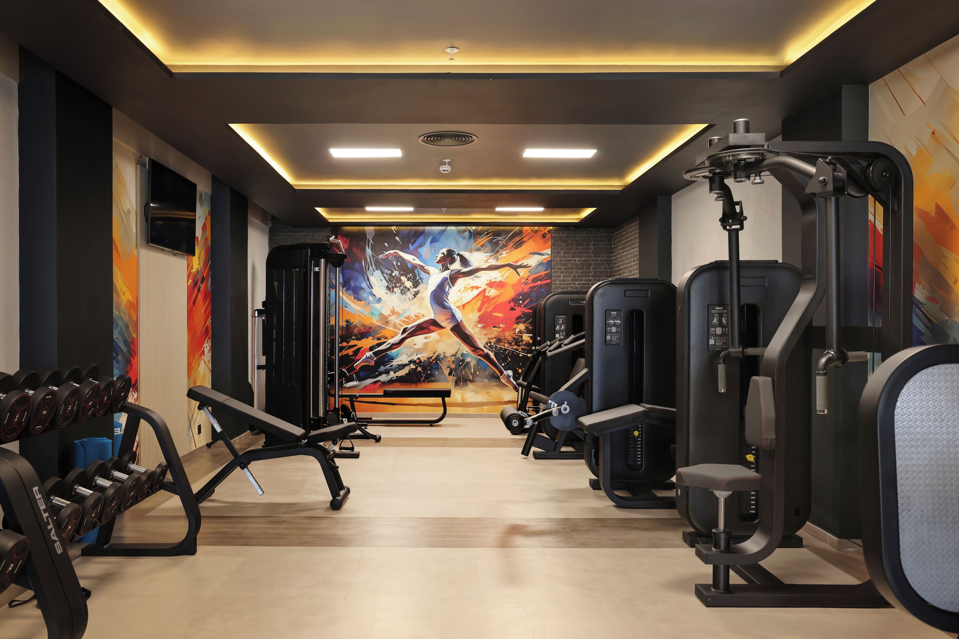 Une salle de sport équipée de matériel de musculation, de bancs et ornée d’une grande fresque murale.