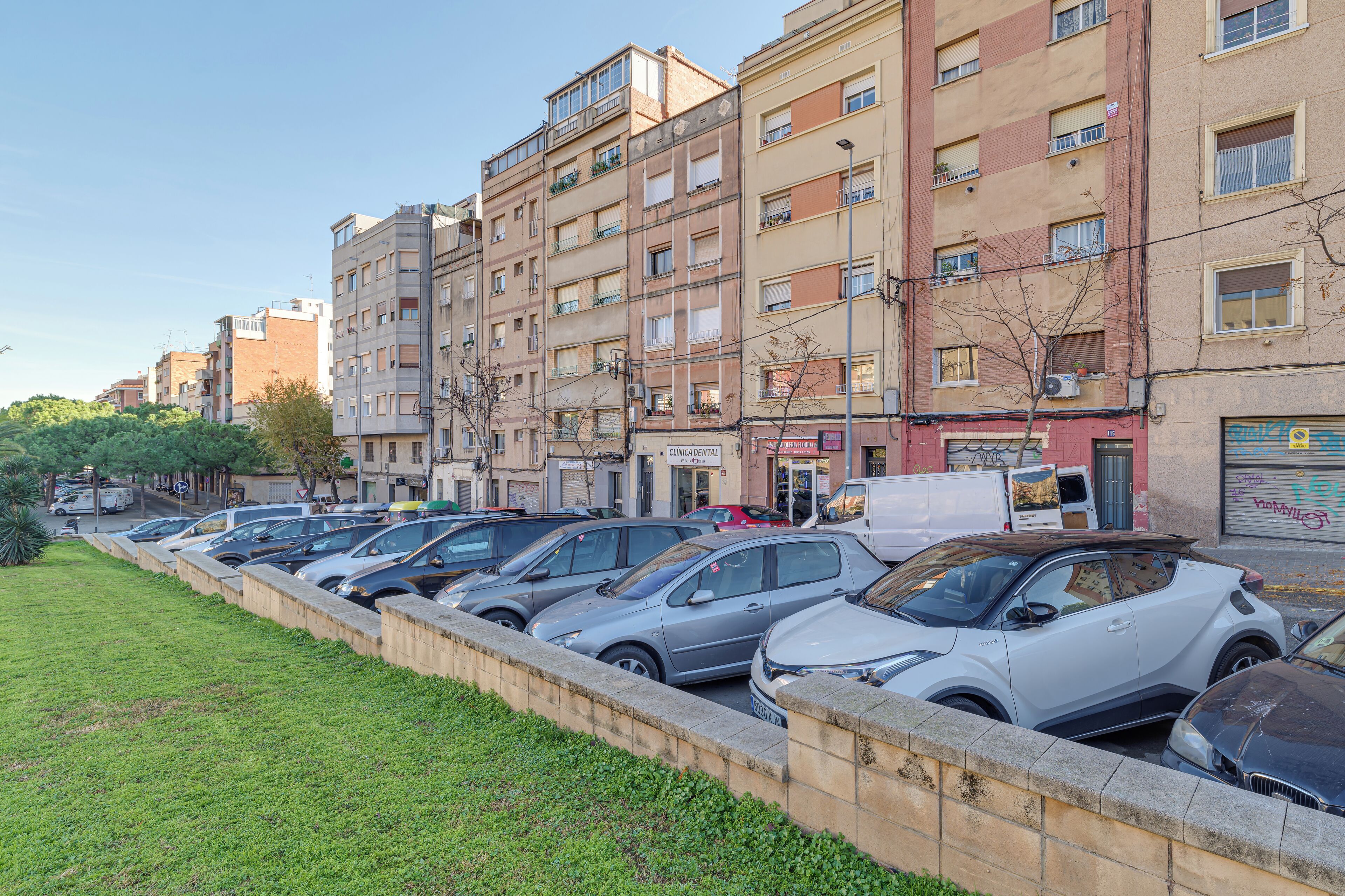 Fila de coches aparcados a lo largo de una acera frente a un edificio de varios pisos con balcones.