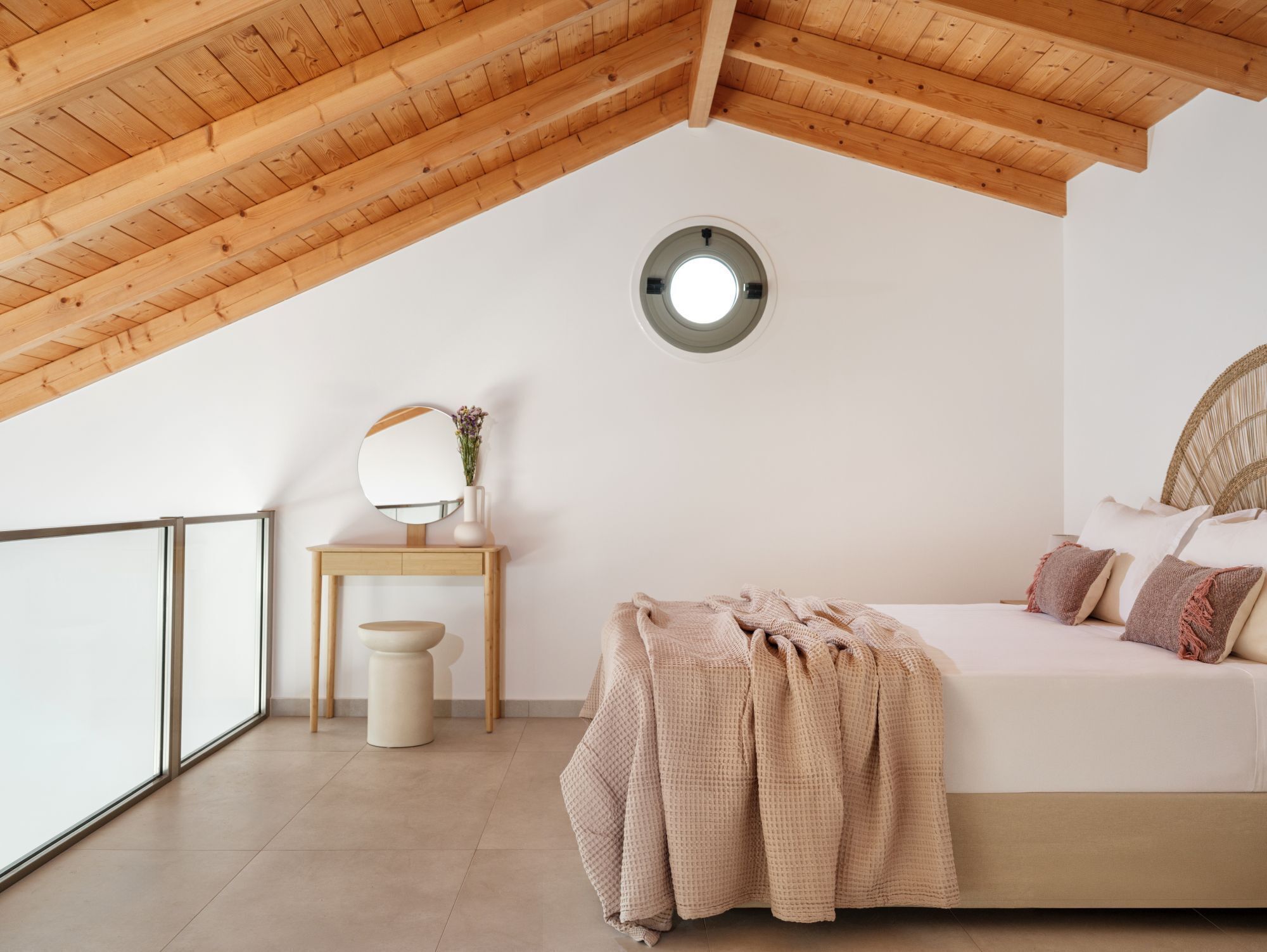 A bedroom with a sloped ceiling, a round mirror, a bedside table, a bed with pillows, and a window.