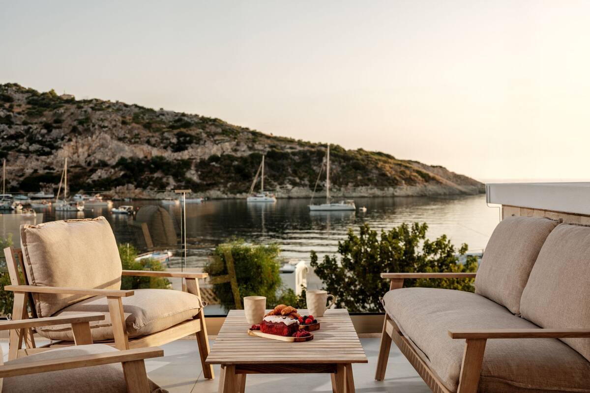 A terrace with wooden furniture, a table with a cake and fruits, and a view of boats and a hilly coastline.