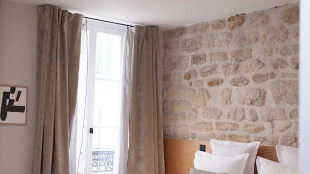 A bedroom with a stone wall, a bed with white bedding, a bench at the foot of the bed, and a window with curtains. - Hotel Tinah: Elegance and Comfort Near the Champs-elysees (Paris)