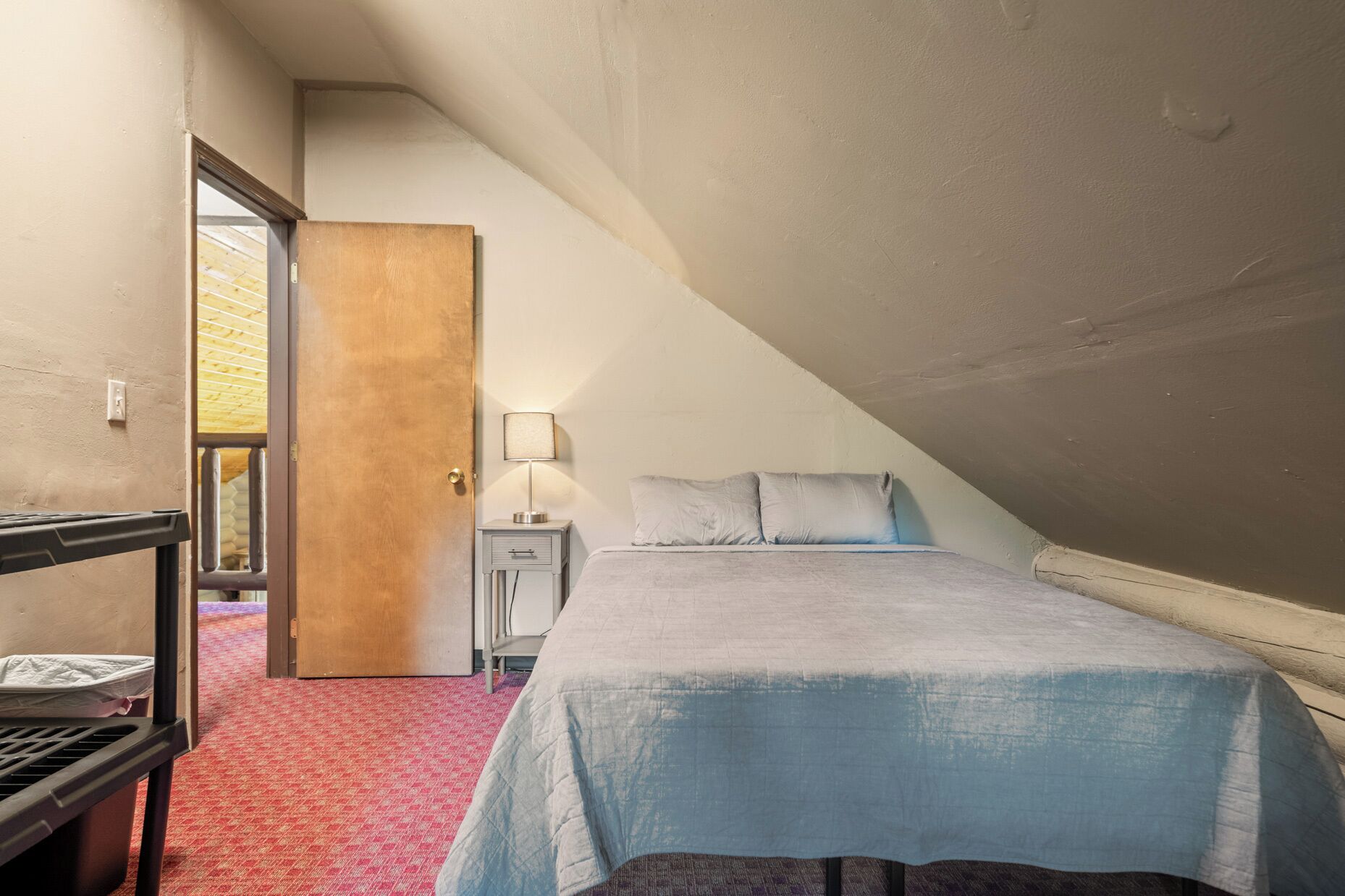 A bedroom with a bed, bedside table, lamp, and a door leading to another room.