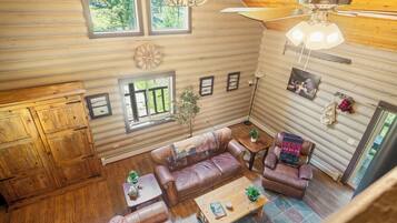 L’intérieur chaleureux d’un chalet, avec un revêtement mural en bois, un ventilateur de plafond, une cheminée et une grande fenêtre donnant sur un paysage verdoyant.